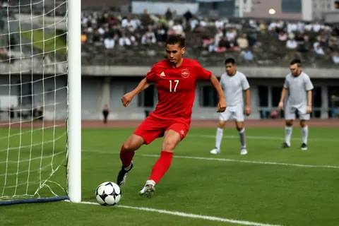 Grounded editorial scene illustrating Kazuyoshi Miura, the world's oldest professional footballer, scored for Fukushima United in a 7-0 victory over Iwaki Furukawa, advancing in the Emperor’s Cup.