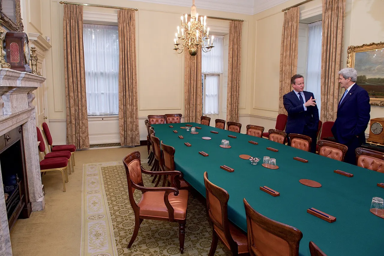 British Prime Minister David Cameron shows U.S. Secretary of State John Kerry the Cabinet Room at 10 Downing Street in London, U.K., with the Prime Minister's armchair angled for easy access, after the two held a bilateral meeting in advance of an anti-corruption summit being hosted by the British leader. [State Department photo/ Public Domain]