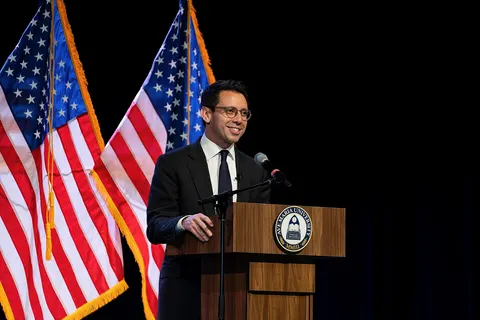 James Fishback delivering remarks at a public event at Ave Maria University in Ave Maria, Florida. The photograph shows him speaking at a podium with United States flags in the background, 2026.