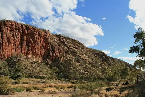 A panoramic view of the western wall of the Glen Helen Gorge in the late afternoon. The Gorge itself can be seen in between peaks on the left. The Finke river, with its mostly dry river bed and glimpses of water, can be seen in the foreground. The trees along the river bed are primarily River Red Gums. The cloud cover is the start of an infrequent dry season cold front which brought rains to the region. This geological formation is part of the West MacDonnell Ranges in Australia's Northern Territory. The layers once lay horizontal at the bottom of the Amadeus basin. The Alice Springs orogeny pushed a 12km thick slab vertical, running roughly east-west for 644km. This gorge is primarily formed of red quartzite, which withstood subsequent erosion better than the softer layers around it. The entire Ranges have been classified as National Park. In the right foreground a couple can be seen enjoying the romantic scene on a bench below the balcony of the Glen Helen camp ground.