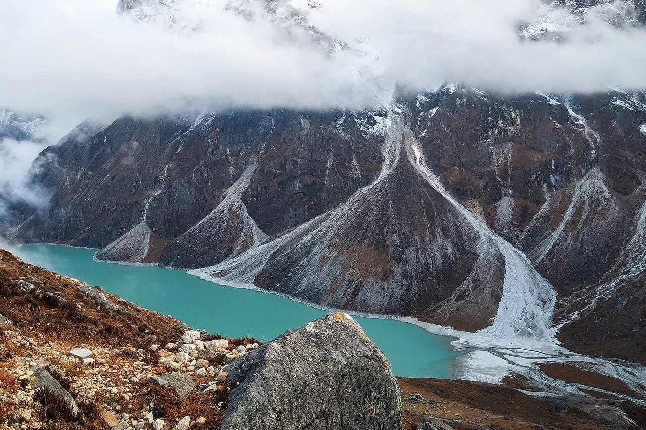 Chola glacial lake (4600) at the base of the north wall of the Cholatse-Taboche mountain massif (6440) with alluvial fans and glacial moraines under a blanket of clouds. Chola Valley, south of the Great Himalayan Range in Mahalangur Himal, Nepal, Himalayas.