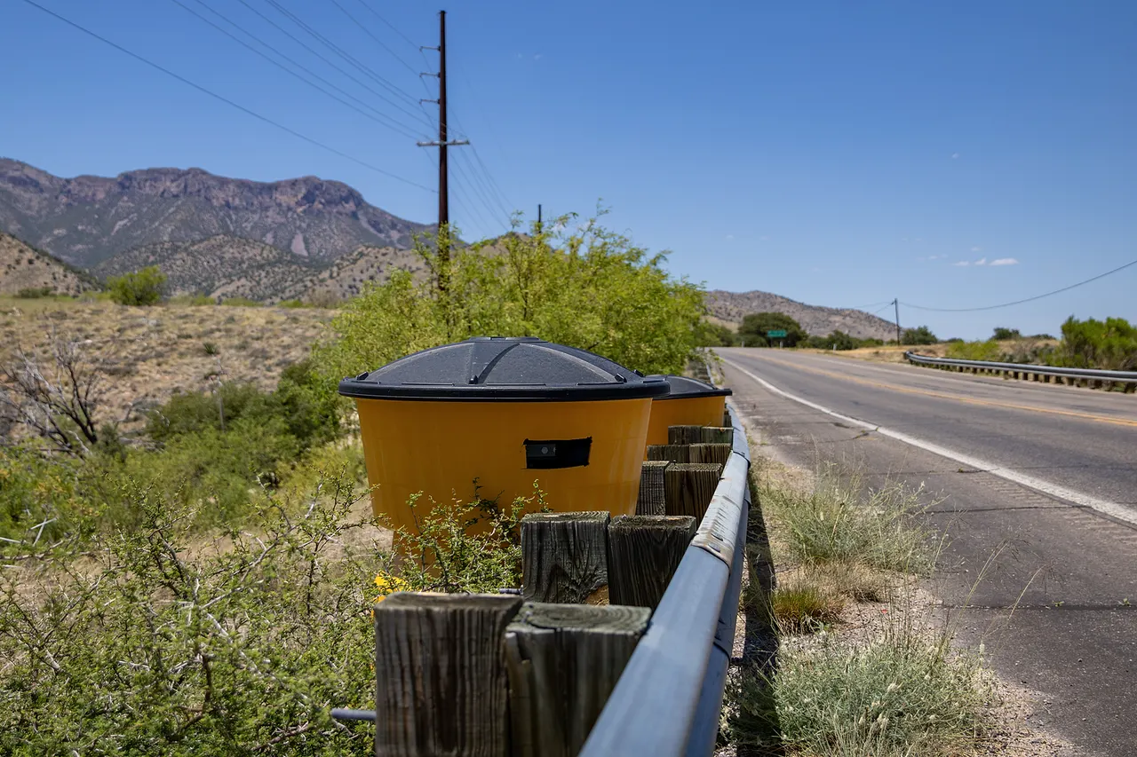 Automated license plate reader (ALPR) system disguised as an impact attenuator sand barrel on the western side of Arizona State Route 92 in Hereford (south of Sierra Vista), Cochise County, Arizona, in May 2024. This unit reads the license plates of vehicles traveling south; another unit on the opposite side of the road reads the plates of vehicles traveling north.